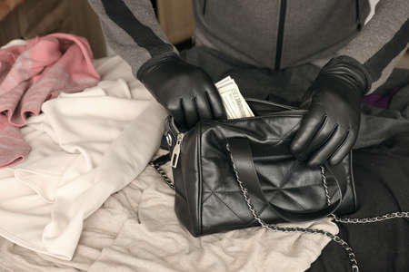 Robber in black outfit and gloves see in opened women bag. The thief takes out US dollar bills from...