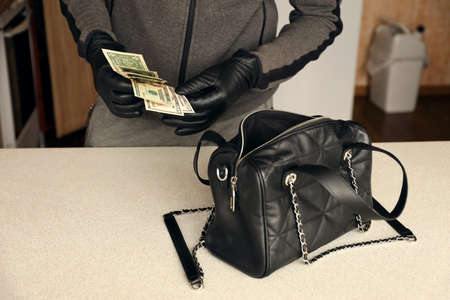 Robber in black outfit and gloves see in opened women bag. The thief takes out US dollar bills from...