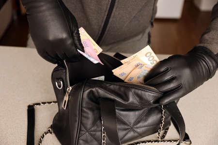 Robber in black outfit and gloves see in opened women bag. The thief takes out the ukrainian money...