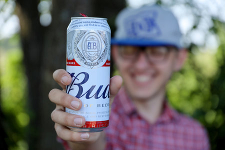 SUMY, UKRAINE - AUGUST 01, 2021: Young man raise Budweiser Bud beer can and shows BUD logo on blurred river and trees background. Budweiser is one of the most popular beer brands in the United Statesのeditorial素材