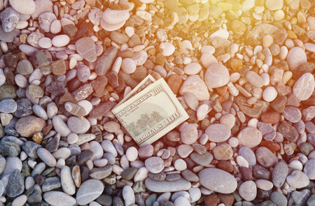 Hundred dollars half covered with round rocks lie on beach close up. Dollar bills partially buried in sea shore stones. Concept finance money holiday relax vacation and money losing in travelの写真素材
