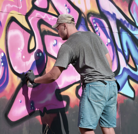 Young caucasian male graffiti artist drawing big street art painting in blue and pink tones. Stylish man in denim shorts and grey t shirt made new graffitti artwork outdoors in bright sunny dayの写真素材