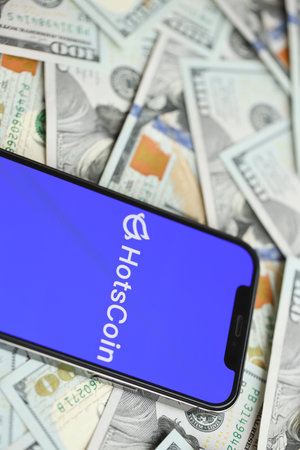 KYIV, UKRAINE - MARCH 15, 2024 Hotscoin logo on iPhone display screen with many hundred dollar bills. Cryptocurrency exchange portalのeditorial素材