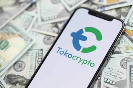 KYIV, UKRAINE - MARCH 15, 2024 Tokocrypto logo on iPhone display screen with many hundred dollar bills. Cryptocurrency exchange portalのeditorial素材