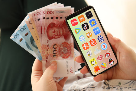 KYIV, UKRAINE - APRIL 1, 2024 Asian apps icon on smartphone screen and money in female hand. iPhone display with app logo and chinese yuan money bills in women hands close upのeditorial素材
