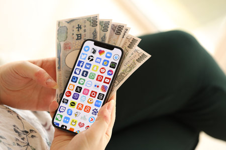 KYIV, UKRAINE - APRIL 1, 2024 Many apps icon on smartphone screen and money in female hand. iPhone display with app logo and japanese yen money bills in women hands close upのeditorial素材