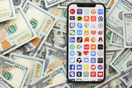 KYIV, UKRAINE - APRIL 1, 2024 Many apps icon on smartphone screen and many usd money bills. iPhone display with app logo and lot of hundred dollars banknotes close upのeditorial素材