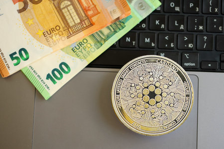 KYIV, UKRAINE - AUGUST 17, 2024 Cardano ADA coin with big amount of european euro bills on laptop surface close upのeditorial素材