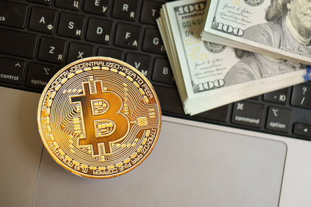 KYIV, UKRAINE - AUGUST 17, 2024 Bitcoin BTC coin with big amount of hundred US dollar bills on laptop surface close upのeditorial素材