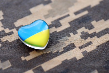 Glass heart with Ukrainian flag lies on military camouflage fabric. Concept of support and defend Ukraineの写真素材