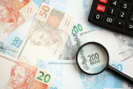 Brazilian reais banknotes and calculator with magnifying glass. The current Brazil real money bills and calc with loupe lies on table in big amount. The concept of taxation and money count in Brazilの写真素材