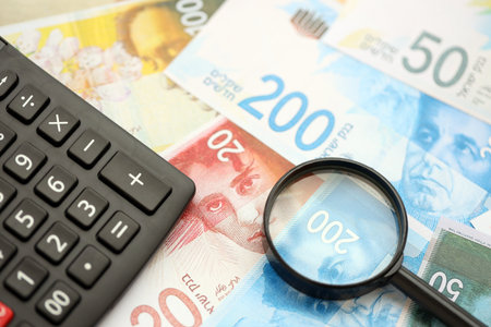 Israel New Shekels banknotes and calculator with magnifying glass. The current Israel money bills and calc with loupe lies on table in big amount. The concept of taxation and money count in Israelの写真素材