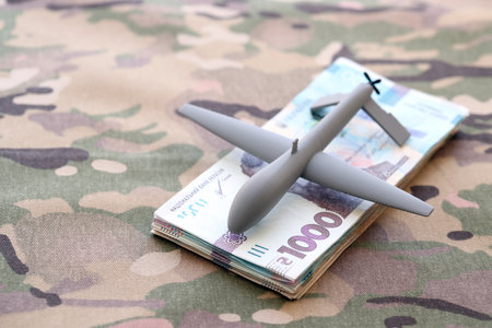 Unnamed grey toy drone and ukrainian money bills on camouflage fabric close up.の写真素材