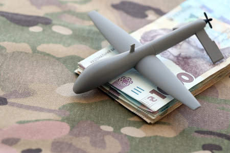 Unnamed grey toy drone model and ukrainian money bills on camouflage fabric close up.