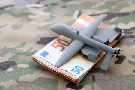 Unnamed grey toy drones and Euro bills stack on camouflage fabric close up. Drone manufacturing fundsの写真素材