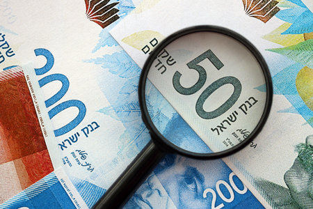 Israel New Shekels banknotes and magnifying glass. The current Israel money bills with loupe lies on table in big amount. The concept of financial monitoring and taxation in Israelの写真素材