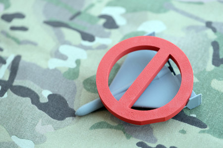 Unnamed grey toy drone and red prohibition sign on camouflage fabric close up. Anti-aircraft warfare...