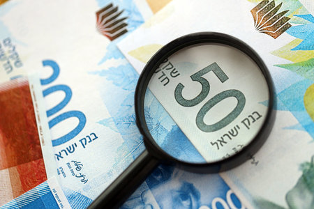 Israel New Shekels banknotes and magnifying glass. The current Israel money bills with loupe lies on table in big amount. The concept of financial monitoring and taxation in Israelの写真素材