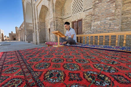 Uzbek man sitting on the carpet and reading in the courtyard of Chor Bakr religious complex, in Bukhara, Uzbekistanのeditorial素材