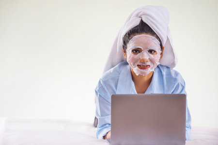 Portrait of young woman applying facial mask on her face during using laptop on bedroomの写真素材