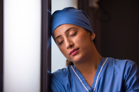 Close-up of a exhausted female nurse standing by window with her eyes closed in hospital.
