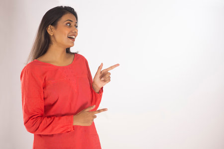 Portrait of a cheerful young woman pointing sideways while standing against white backgroundの写真素材