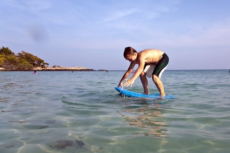 boy is surfing on a small surfboard in a beautiful sea with crystal clear water and blue sky, his brother holds the surfboard on his backの写真素材