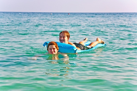 brothers are playing together in a beautiful sea on an air mattress with crystal clear water and blue skyの写真素材