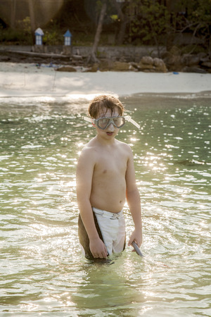 young boy wearing a diving mask is walking at the beautiful beach, enjoying the waterの写真素材
