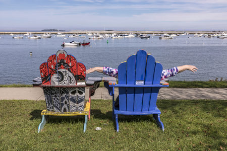 PLYMOUTH, USA - SEP 26, 2017: colorful painted beach chairs by local artists at the promenade of Plymouth with harbor and ships in background.のeditorial素材