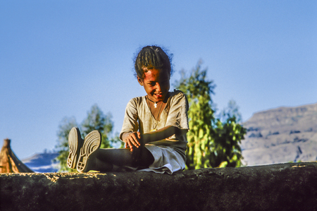 LALIBELA, ETHOPIA - JULY 1, 1998: young girl mith plastic boots sitting at a loam wall enjoys reading a funny text. Girl wears a coptic cross as symbol for her religion.のeditorial素材