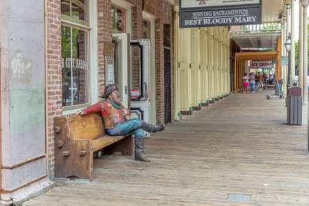 Sacramento, USA - June 4, 2022: wooden statue of cowboy sitten on a bench in the old town of Sacramento with smoking pipe in front of a bar and tlurists in background at historic sidewalk.のeditorial素材