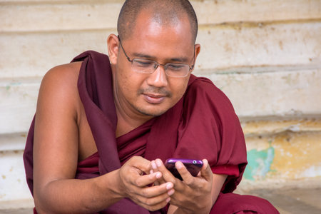 Pagan, Myanmar - August 14, 2015: Monk in traditional dress uses a mobile or smart phone to get...