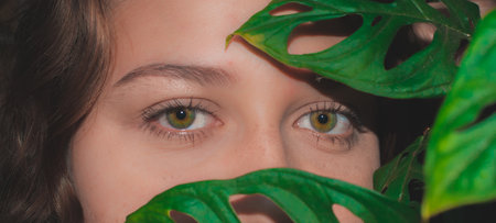 Close up portrait of a young woman with green eyes and monstera leaf. face of a beautiful young woman covering her face with leaves. portrait of woman with natural makeupの写真素材