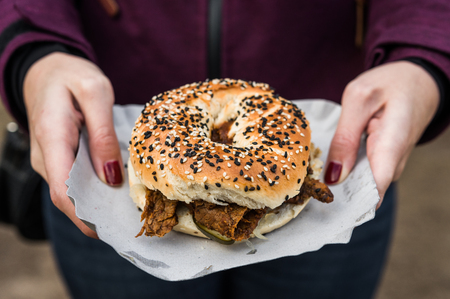 Close up photo of a bagel with sesame seeds, pastrami beef meat, pickles, and mustard at a street...