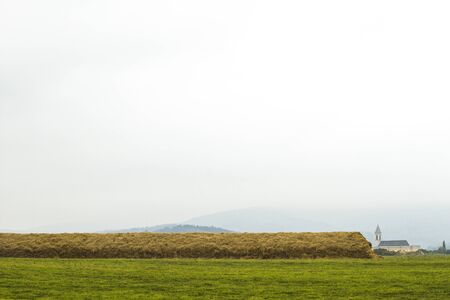 Rural landscape background. Beautiful, cozy, scenic panorama, vertical composition. Meadow and very long haystack, Catholic church and hills in the fog. Foggy weather, early autumn. Czech Republic.の写真素材