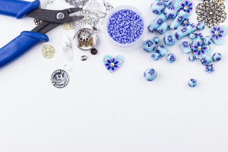Jewelry making and beading. Seedbeads, metal components, polyclay purple blue flower beads mix, crimp pliers on white background. Selective focus. Top view.の写真素材