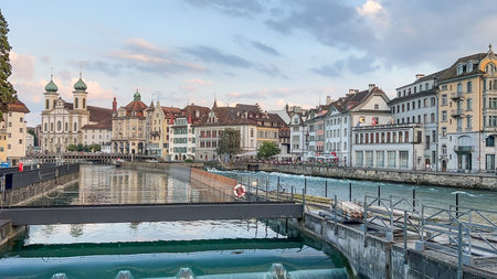 A scenic view of Lucerne, Switzerland, featuring the River Reuss, historic buildings, and a cloudy sky. The Jesuit Church stands prominently on the left.の写真素材