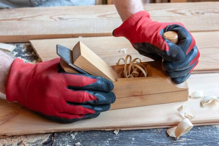 A carpenter in red cloth gloves holds a wooden jointer in his hands close-up. Sawdust, shavings, boards on the tableの写真素材