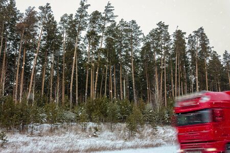 The road in a pine forest. Winter, it is snowing. Red truck rides