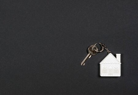 A keychain in the form of a white house and a small metal key on a black background. The concept of buying, selling, renting real estate, mortgages, neighbors, your homeの写真素材