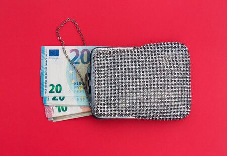Banknotes of 20 and 10 euros in a shiny vintage handbag with rhinestones on a red background. The...