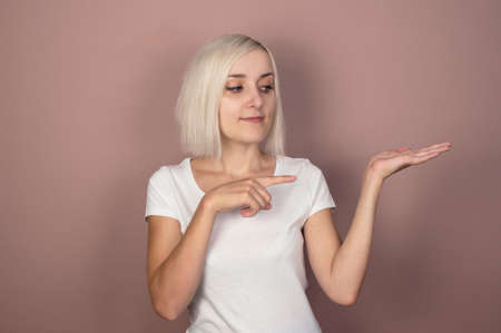 Beautiful young blonde woman in a white T-shirt on a pink background shows a finger. Place for text,...