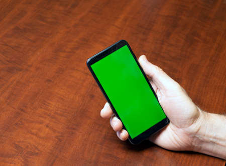 Mobile phone with a green screen in a man's hand on the background of a wooden table. Blank screen for content integrationの写真素材