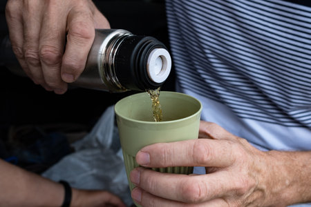 Male hands pour coffee or tea from a flask into a reusable glass. Zero waste concept, outdoor...
