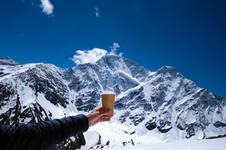 Close-up of a man's hand with a disposable cup against the backdrop of snow-capped mountains and blue sky. Vacation in the mountains, hike, travel. With space for textの写真素材
