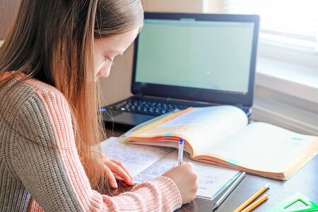 Girl sitting at desk doing lessons with notebook near laptop.