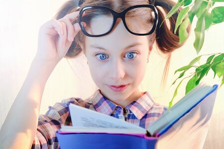 The schoolgirl looks in surprise at the book with big eyes, raising his glassesの写真素材