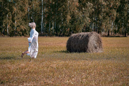 A girl in a white dress runs across the field to a haystackの写真素材