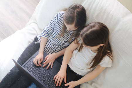 Two school children sitting together on the sofa and typing on a laptopの写真素材